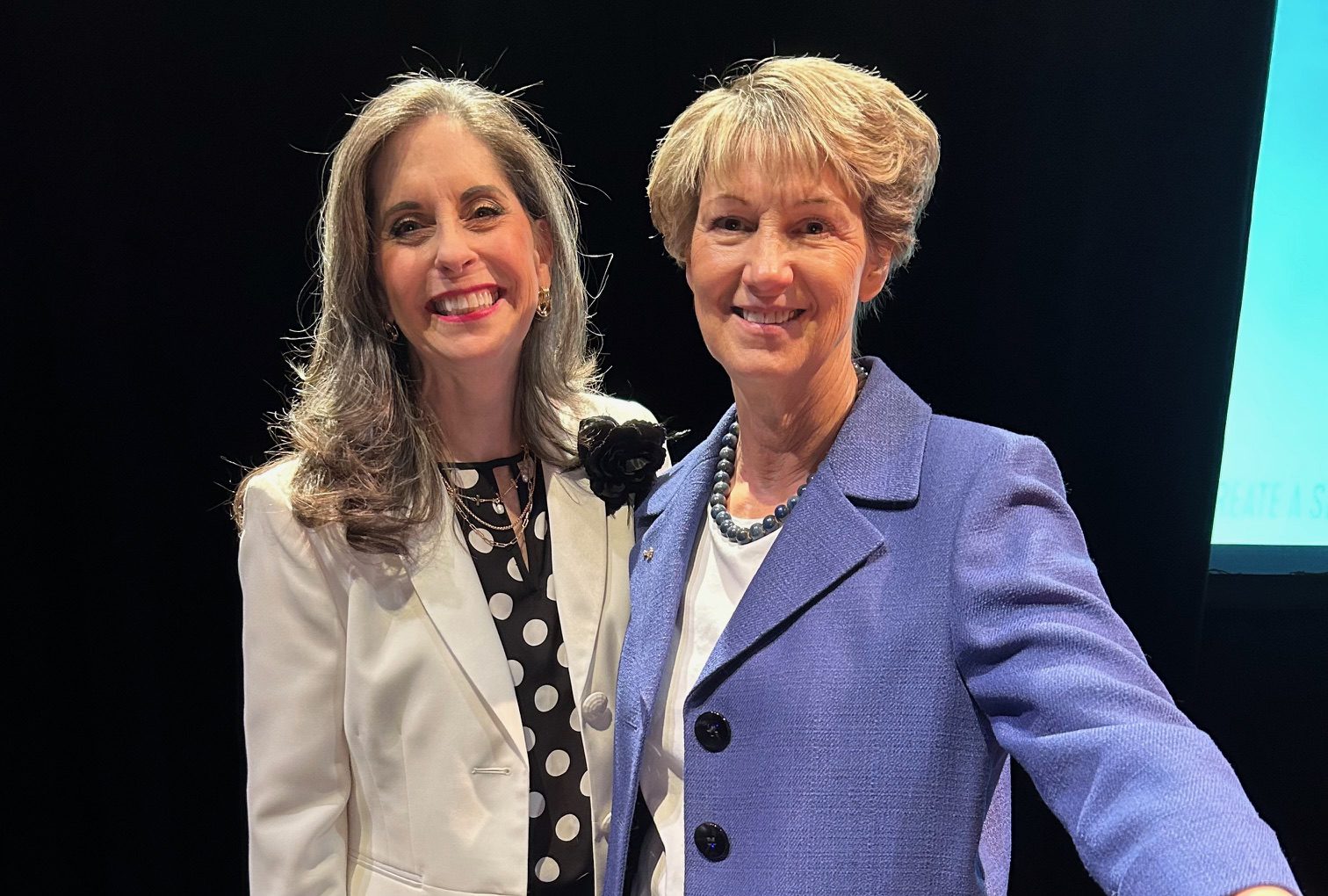 Two women smiling, one in a white jacket and polka dot top, the other in a blue blazer.