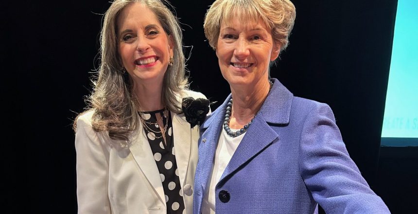 Two women smiling, one in a white jacket and polka dot top, the other in a blue blazer.